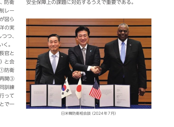 日米韓防衛相会談で握手する3人の男性、背景には木目調の壁と書類が置かれている。